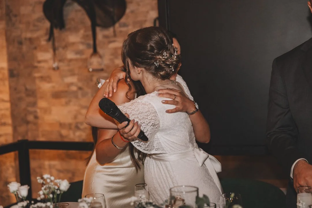 Two women hugging at a celebration, one holding a microphone, with a brick wall and flowers in the background.