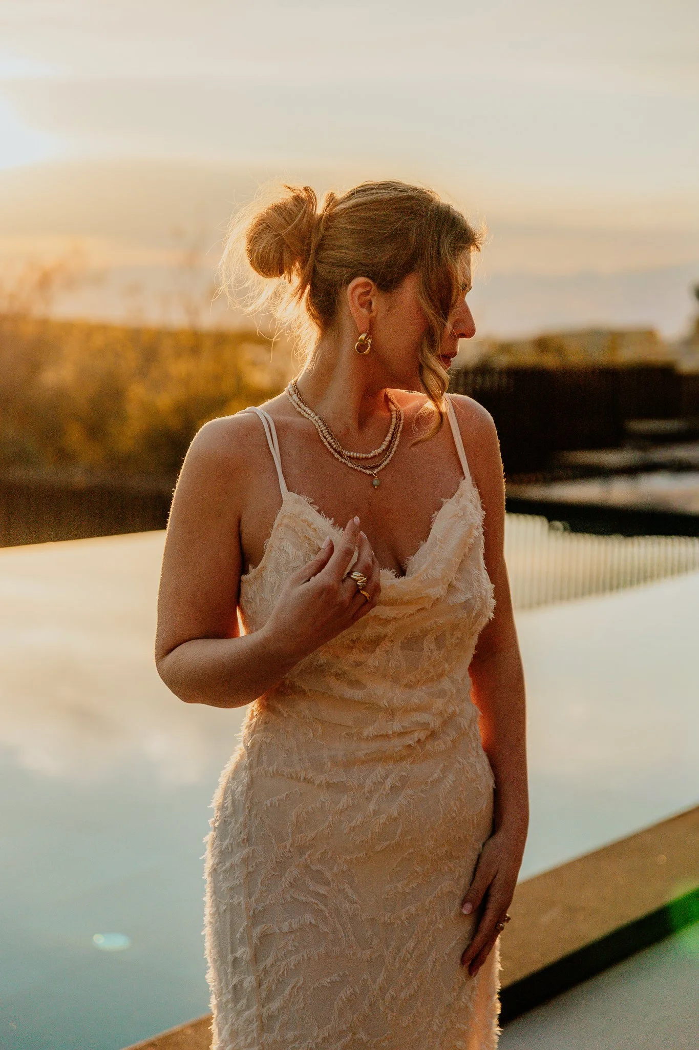 A woman in a beige dress with textured fabric stands near a body of water during sunset at San Canzian, Croatia. Her hair is in a loose bun, and she is wearing earrings, a necklace, and rings. She looks down with a calm expression
