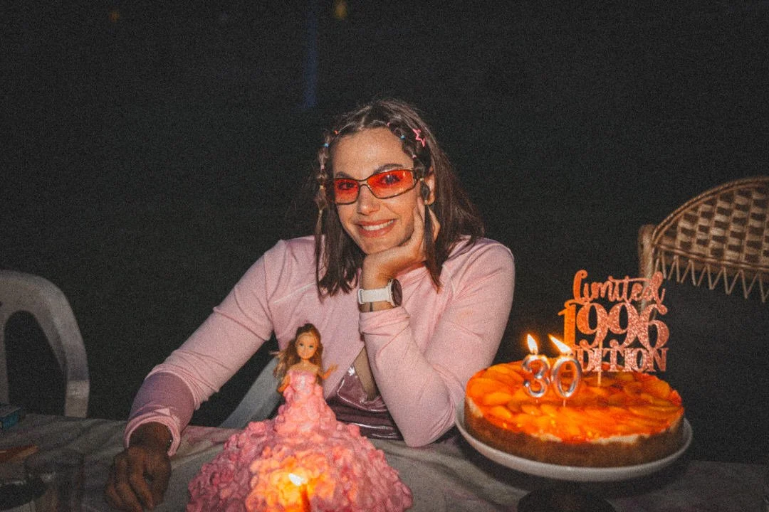 A woman wearing red sunglasses and a pink blazer sitting at a table with birthday treats, including a pink cake with a doll figure on top and a pie with a candle, celebrating a 30th birthday at night. 1990s vibes