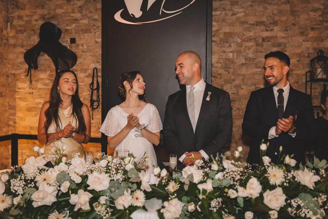 A wedding reception with four people standing behind a table decorated with white flowers, with a woman in a white dress and a man in a black suit in the center, surrounded by two women in black and gold dresses, one of whom is clapping, all smiling.