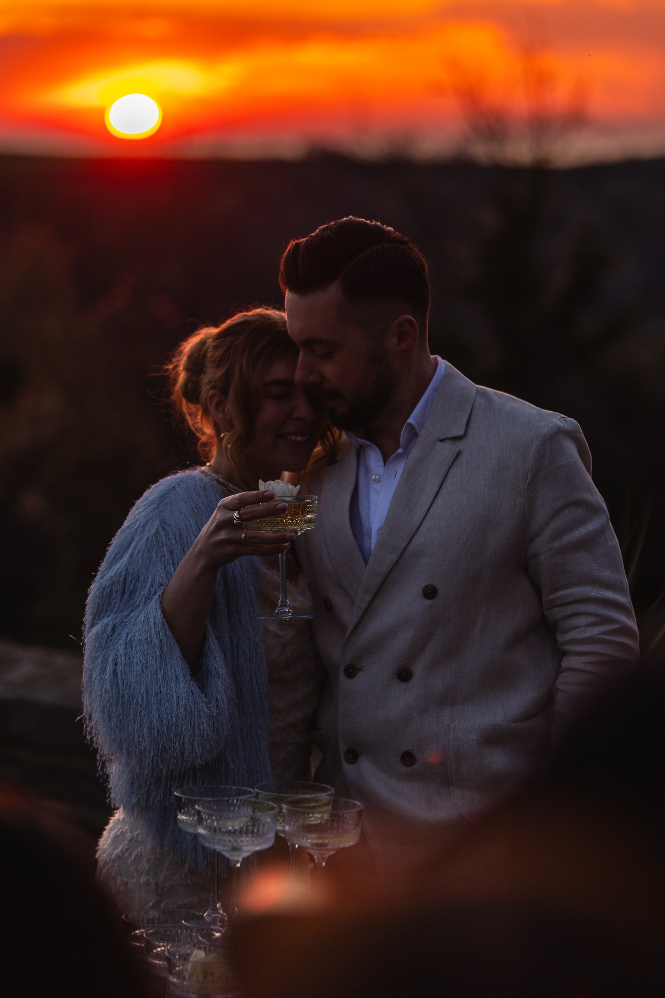 A romantic couple dressed in formal attire, standing close together during a sunset, holding a glass of wine, with several other glasses on a table in front of them.