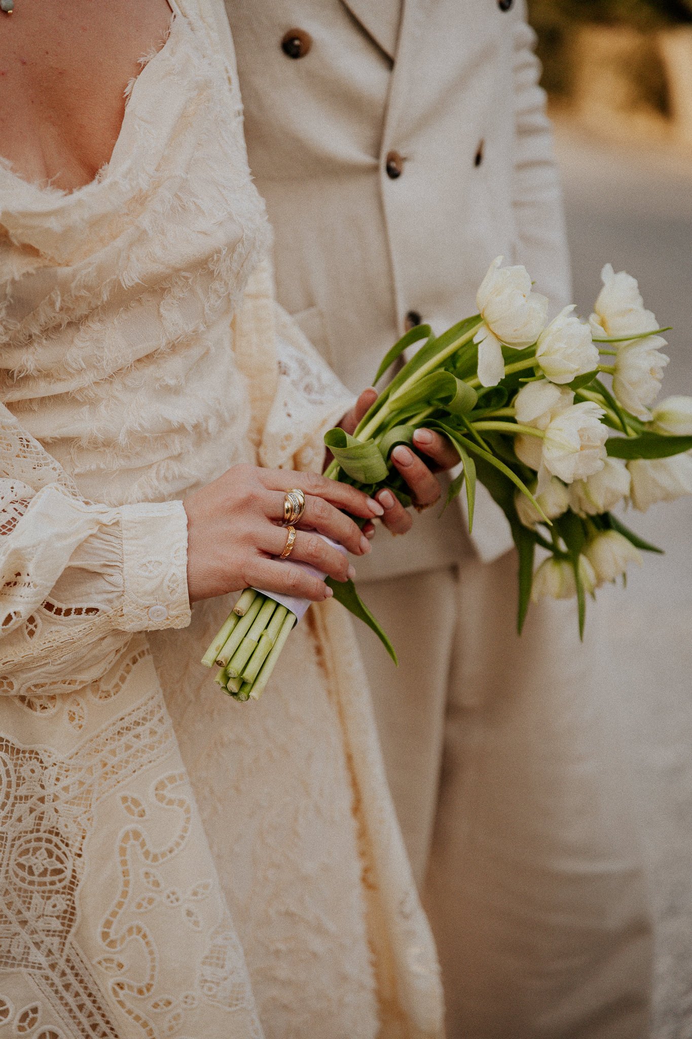 Close-up of a bride and groom holding a bouquet of white tulips during a wedding ceremony, with the bride wearing a lace dress and the groom in a beige suit.