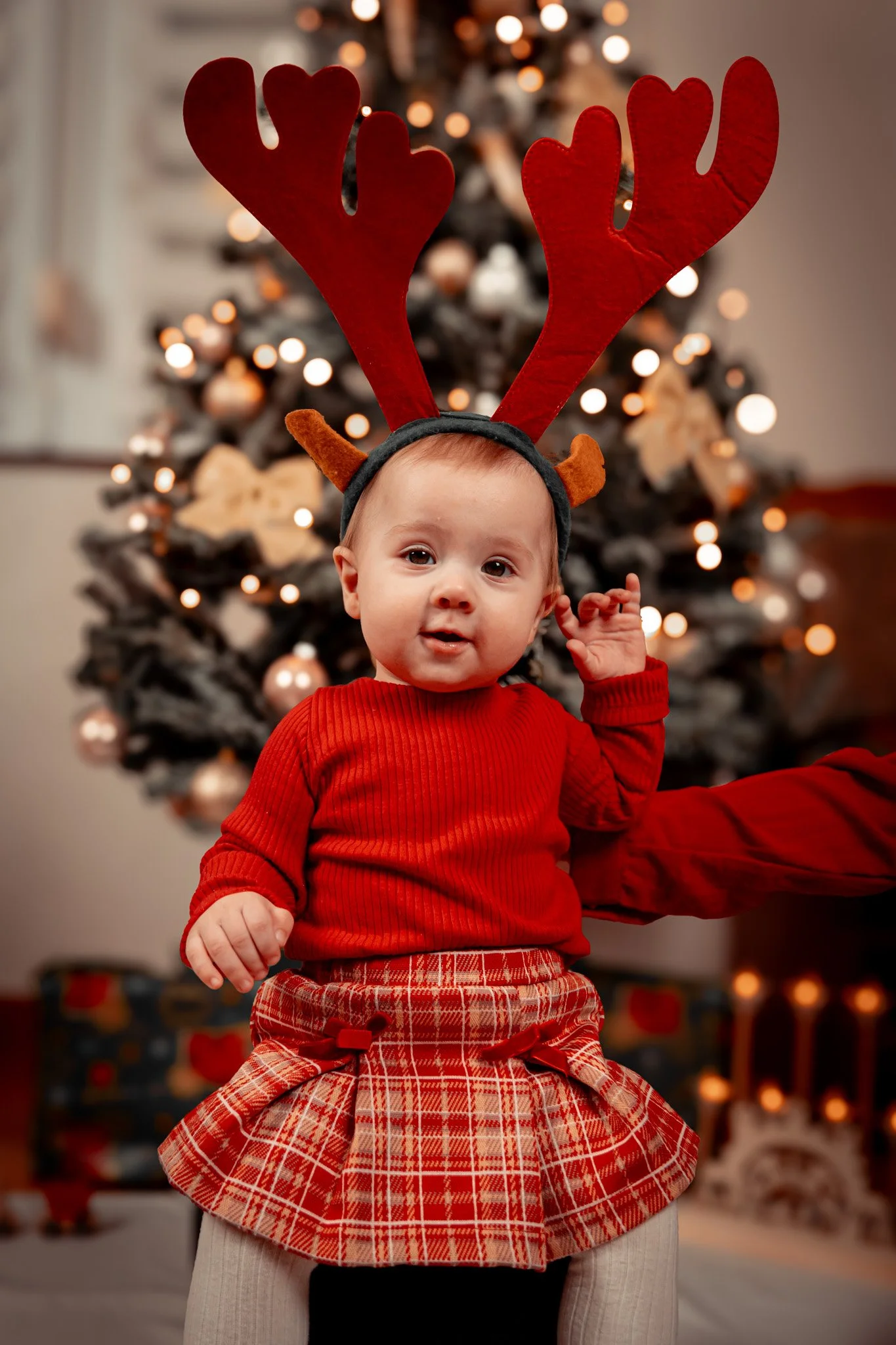 A baby dressed in a red sweater and plaid skirt, wearing reindeer antlers headband, sitting on an adult's lap with a Christmas tree in the background decorated with warm lights.