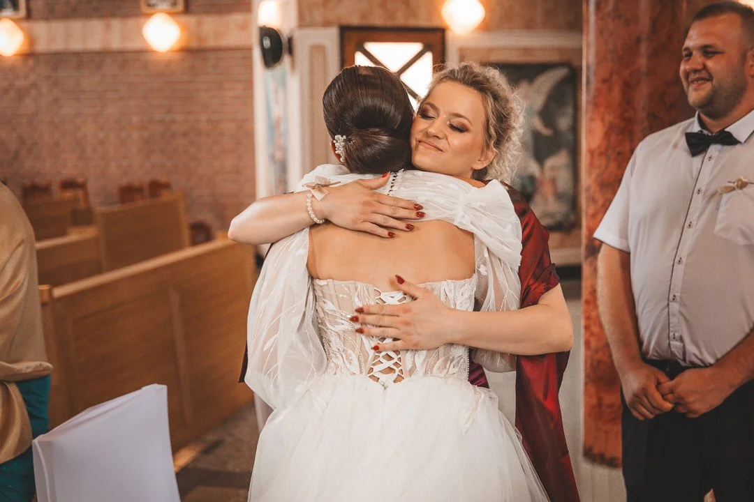 Two women hugging at a wedding reception, one in a white wedding dress and the other in a dark red dress, with a man in a white shirt and black bow tie smiling in the background.