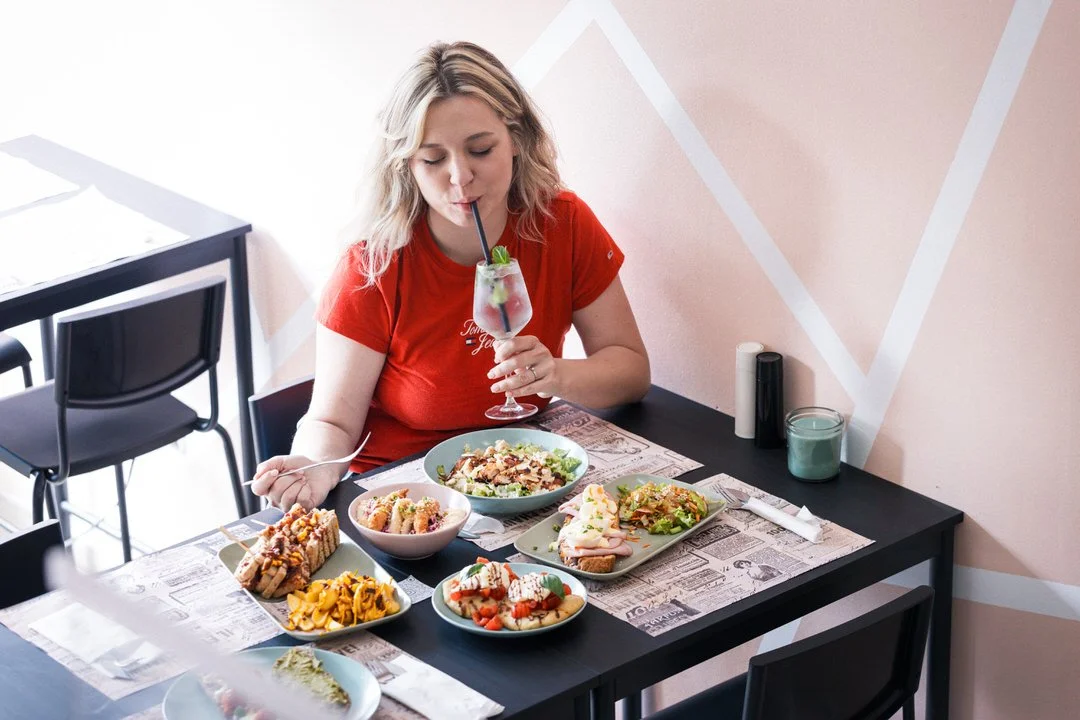 A woman with blonde hair wearing a red shirt drinking a cocktail at a dinner table with various dishes including salads, sandwiches, pasta, and a dessert in a modern restaurant with light pink walls.