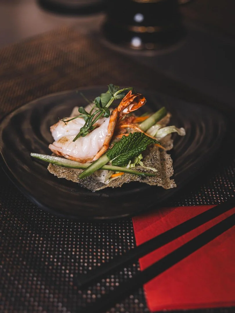 A plate of Asian cuisine featuring a shrimp, garnished with greens and vegetables, served on a black plate with chopsticks nearby.