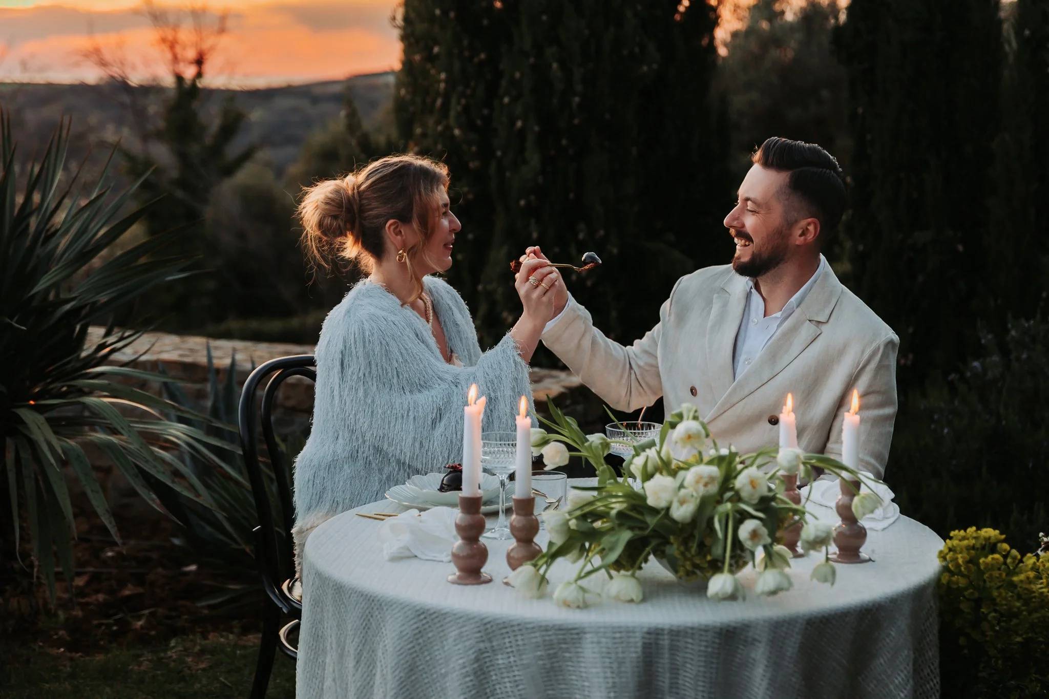 A man and woman enjoying a romantic outdoor dinner at sunset during their San Canzian wedding celebration, with the woman feeding the man a piece of food, surrounded by candles and floral arrangements on the table.