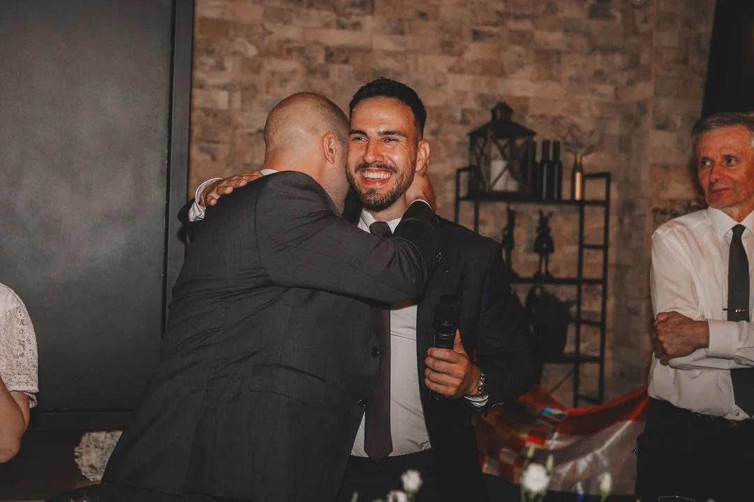 Two men in suits sharing a hug at a social event, with other guests and decorative shelves in the background.