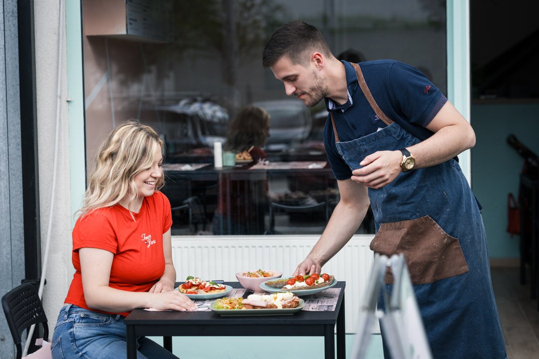 A smiling woman in a red shirt is sitting at an outdoor table with three plates of food, while a man in an apron serves her. They appear to be at a brunch place in Zagreb Croatia