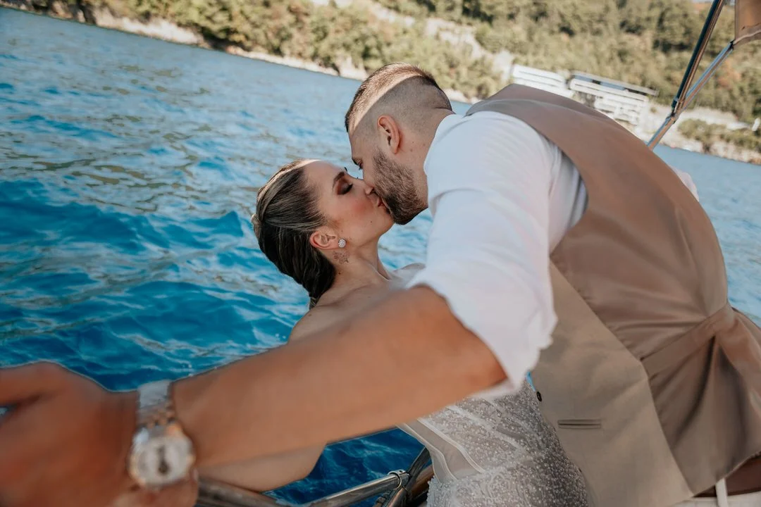 A newlywed couple sharing a kiss on a boat with water and a green shoreline in the background.
