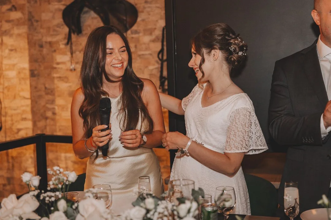 Two women in white dresses at a wedding reception, smiling and talking to each other, with glasses and floral decorations on the table in front of them.