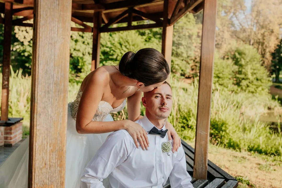Bride leaning over groom, sitting on a park bench, under a wooden pavilion in a lush green park, during daytime.