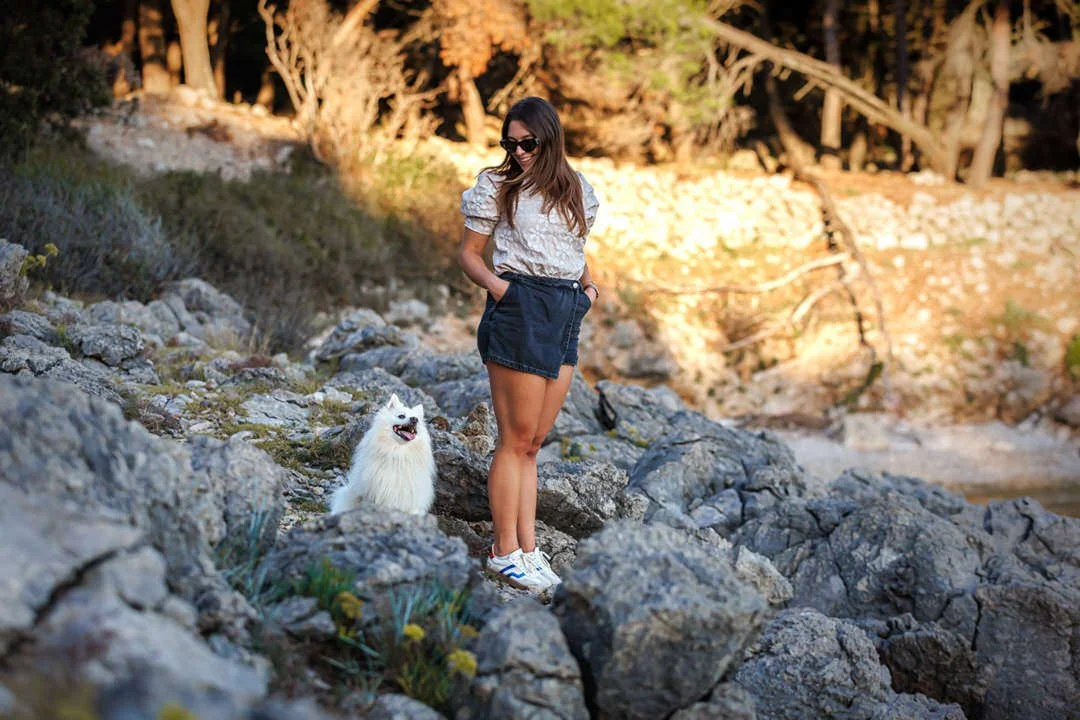 A woman wearing sunglasses, a beige top, black shorts, and white sneakers standing on rocky terrain with a white fluffy dog nearby on Prniba, Krk Island, Croatia
