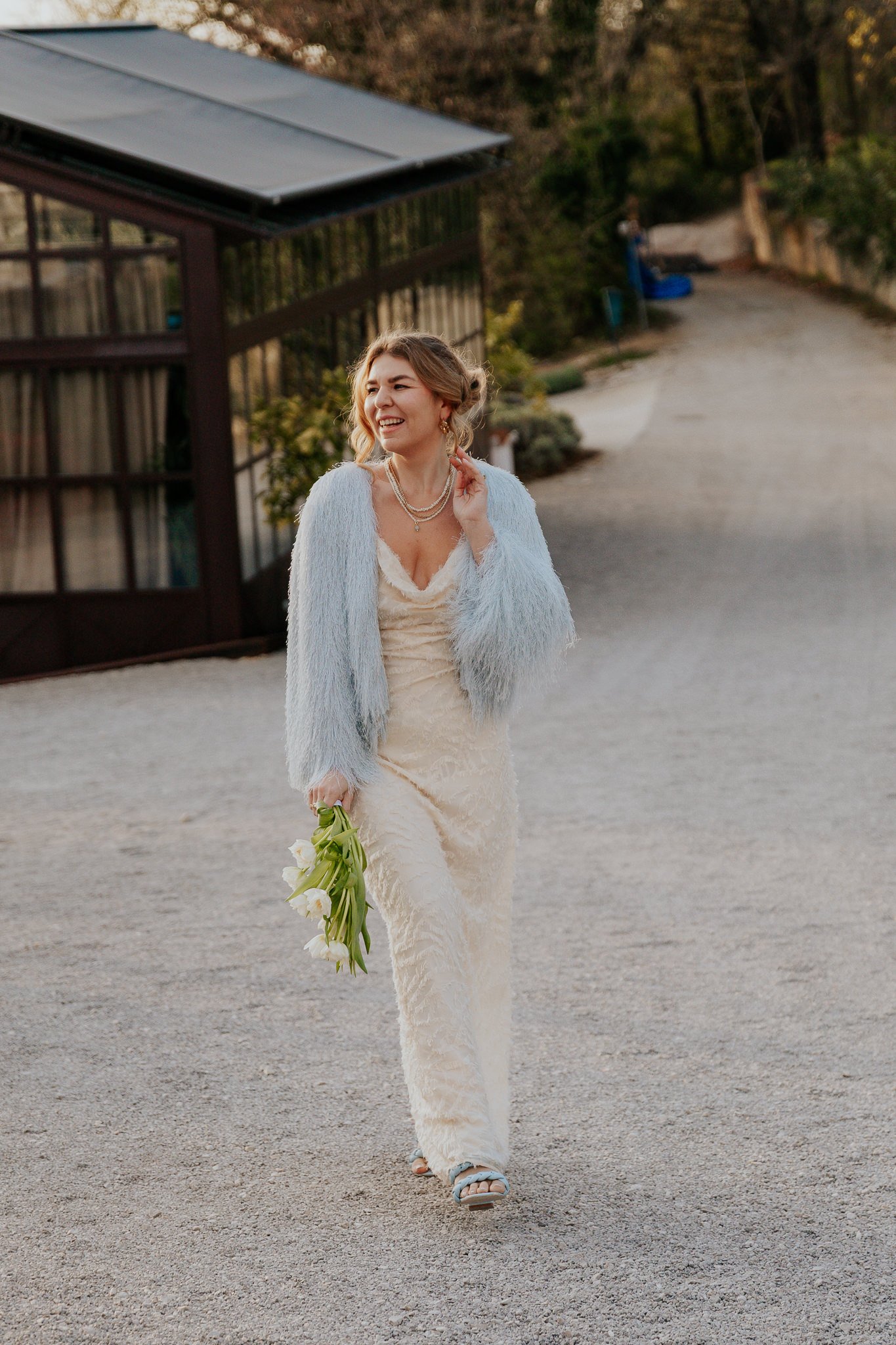 A bride dressed in a long, elegant white dress and a light blue fuzzy jacket is smiling and walking outdoors on a gravel path, holding a small bouquet of white flowers in her right hand.