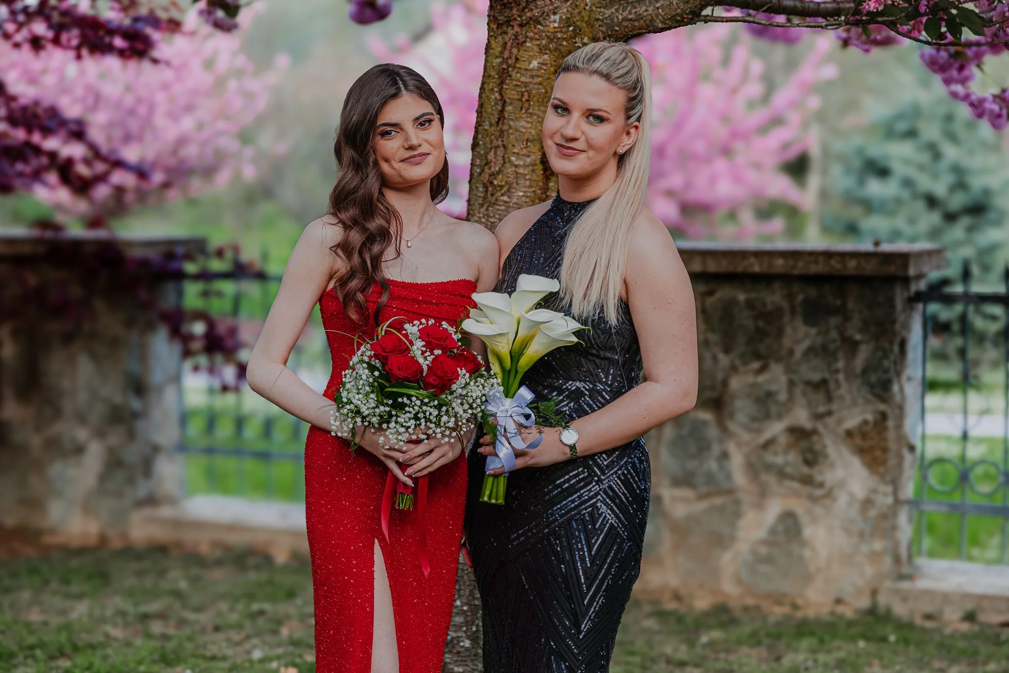 Two women stand outdoors near a tree with pink blossoms, holding bouquets of flowers, one with a red dress and the other with a black dress.