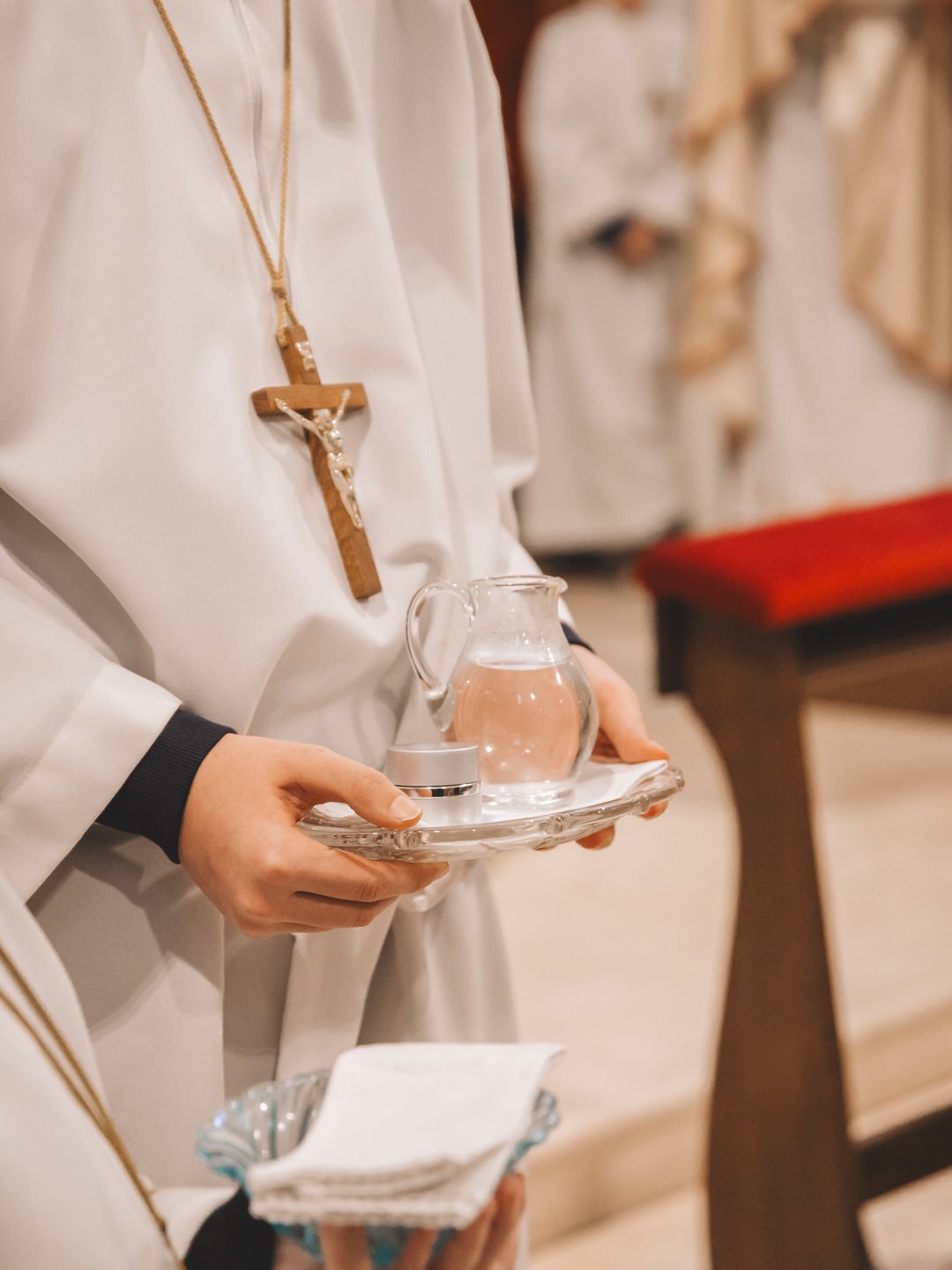 Person in white robe holding a tray with a glass pitcher of water and a small container, wearing a wooden cross necklace in a church setting.