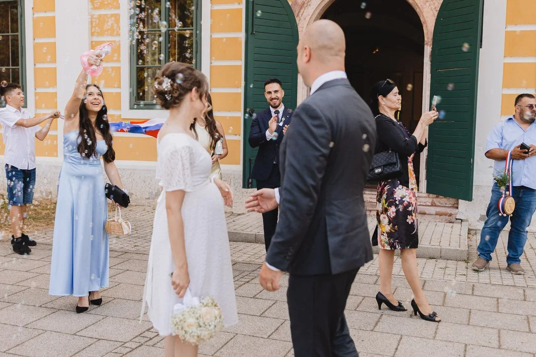 A wedding celebration outside a yellow building with green shutters, featuring a bride in a white dress holding a bouquet, a groom in a dark suit, and guests celebrating with bubbles and medals.