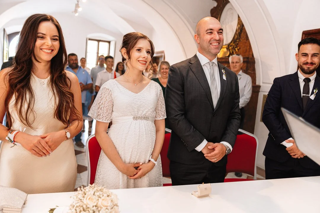 People attending a wedding ceremony indoors, standing in front of a table with floral decorations, with the bride and groom at the center, smiling.