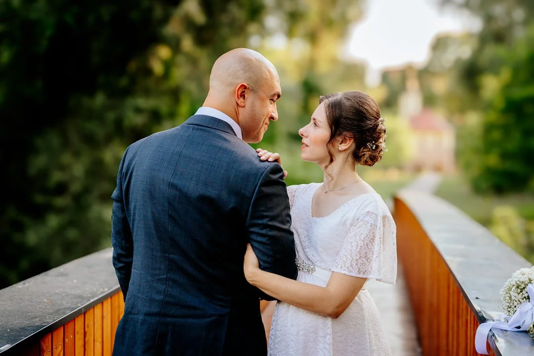 A bride and groom standing face to face on a bridge outdoors, gazing into each other's eyes, with lush green trees in the background.
