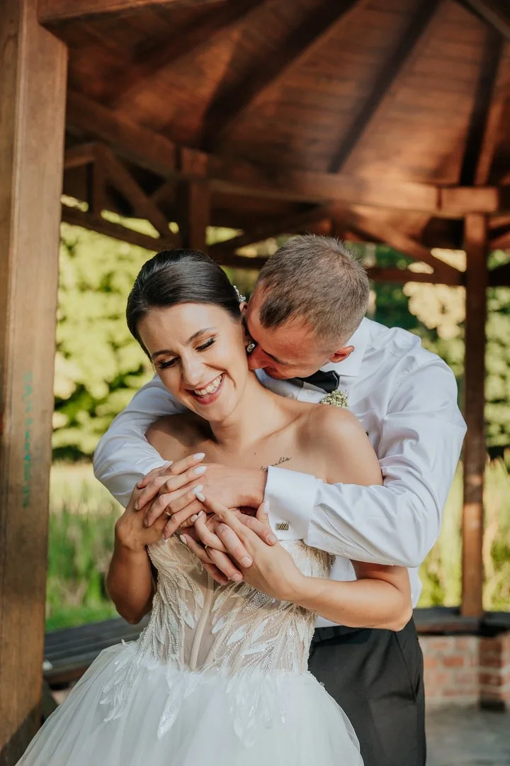 A bride and groom sharing a joyful moment, with the groom hugging the bride from behind and both smiling, outdoors under a wooden pavilion.