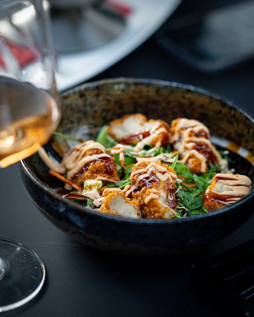 Close-up of a black bowl containing crispy fried appetizer pieces topped with drizzles of creamy sauce, served on a bed of greens, with a glass of white wine nearby in Aera restaurant in Bobingen Germany