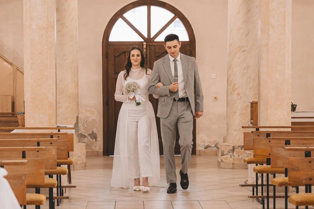A bride and groom walking down the aisle inside a church in Slavonia Croatia, with the bride holding a bouquet of white flowers and the groom holding a microphone.
