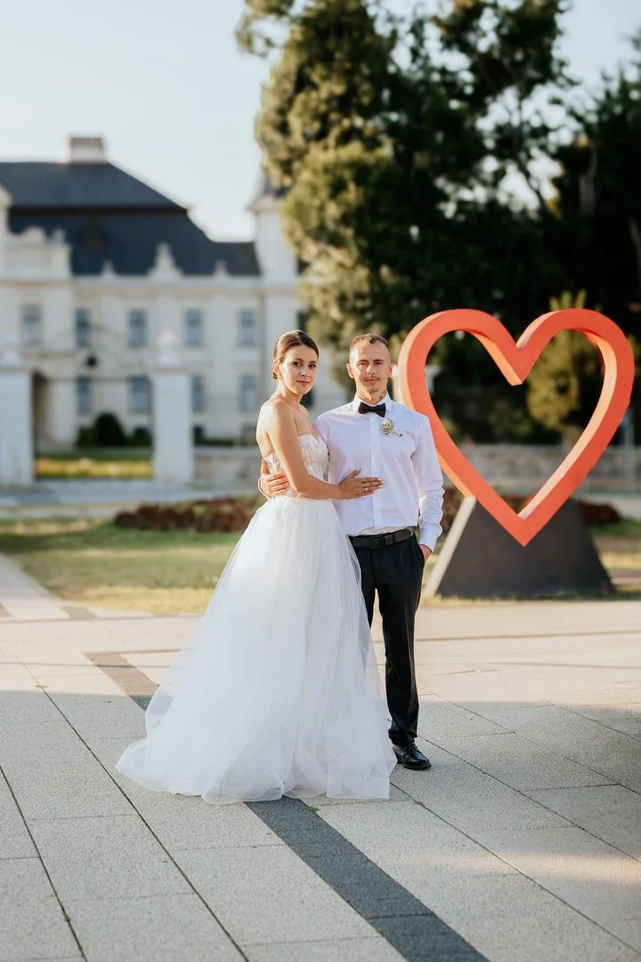 A bride and groom standing together outdoors, with a large red heart sculpture behind them, on a paved area near a large white building and trees.
