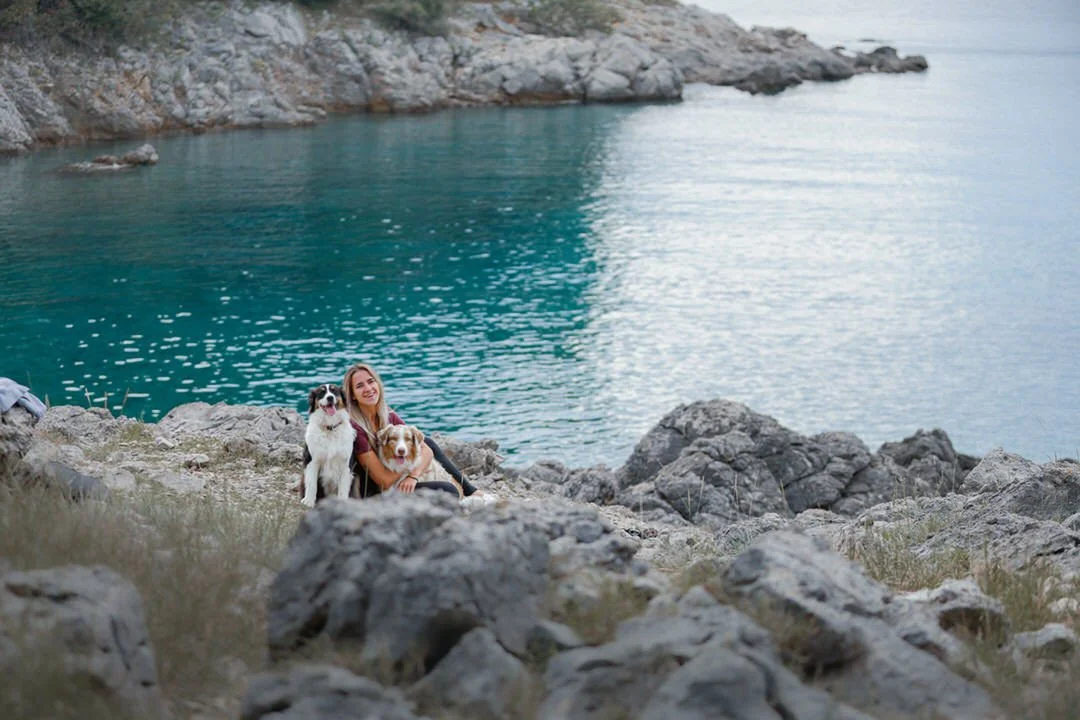 A woman sitting on rocky terrain next to two dogs, with a body of water and rocky cliffs in the background in Prniba, Krk Island, Croatia.