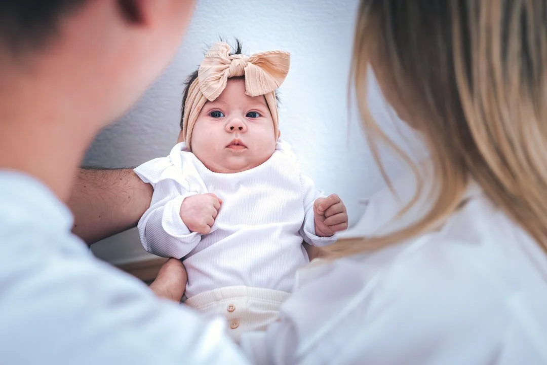 A baby girl with a beige headband and bow, lying down between her parents, looking at the camera.