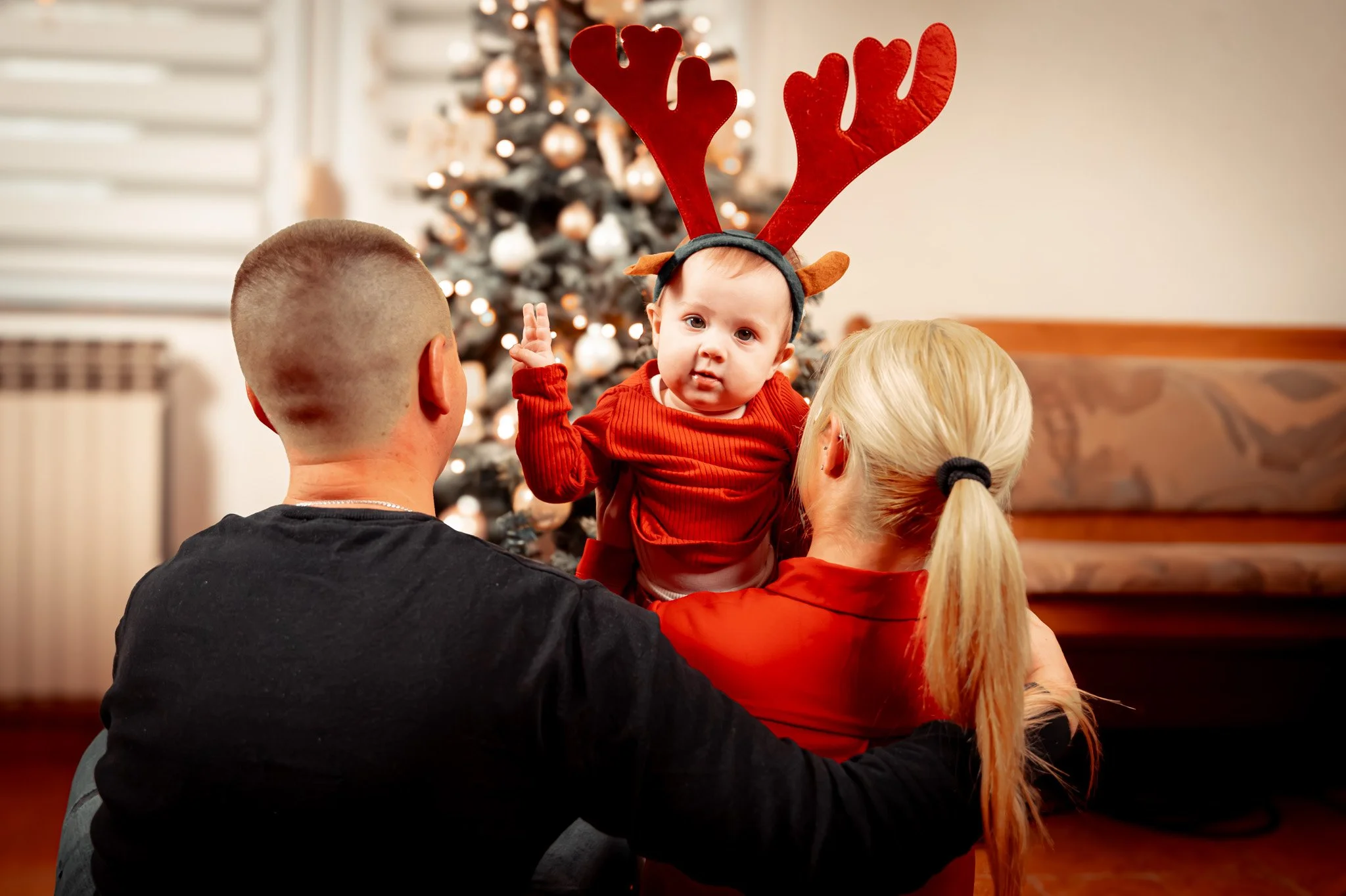 A baby wearing reindeer antlers being held by two adults, with a decorated Christmas tree in the background.