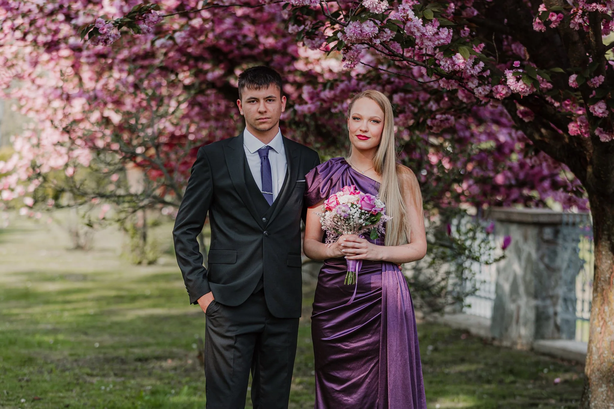 A man in a black suit and a woman in a purple dress standing under a blooming pink tree, holding a bouquet of flowers at prom session in Orahovica