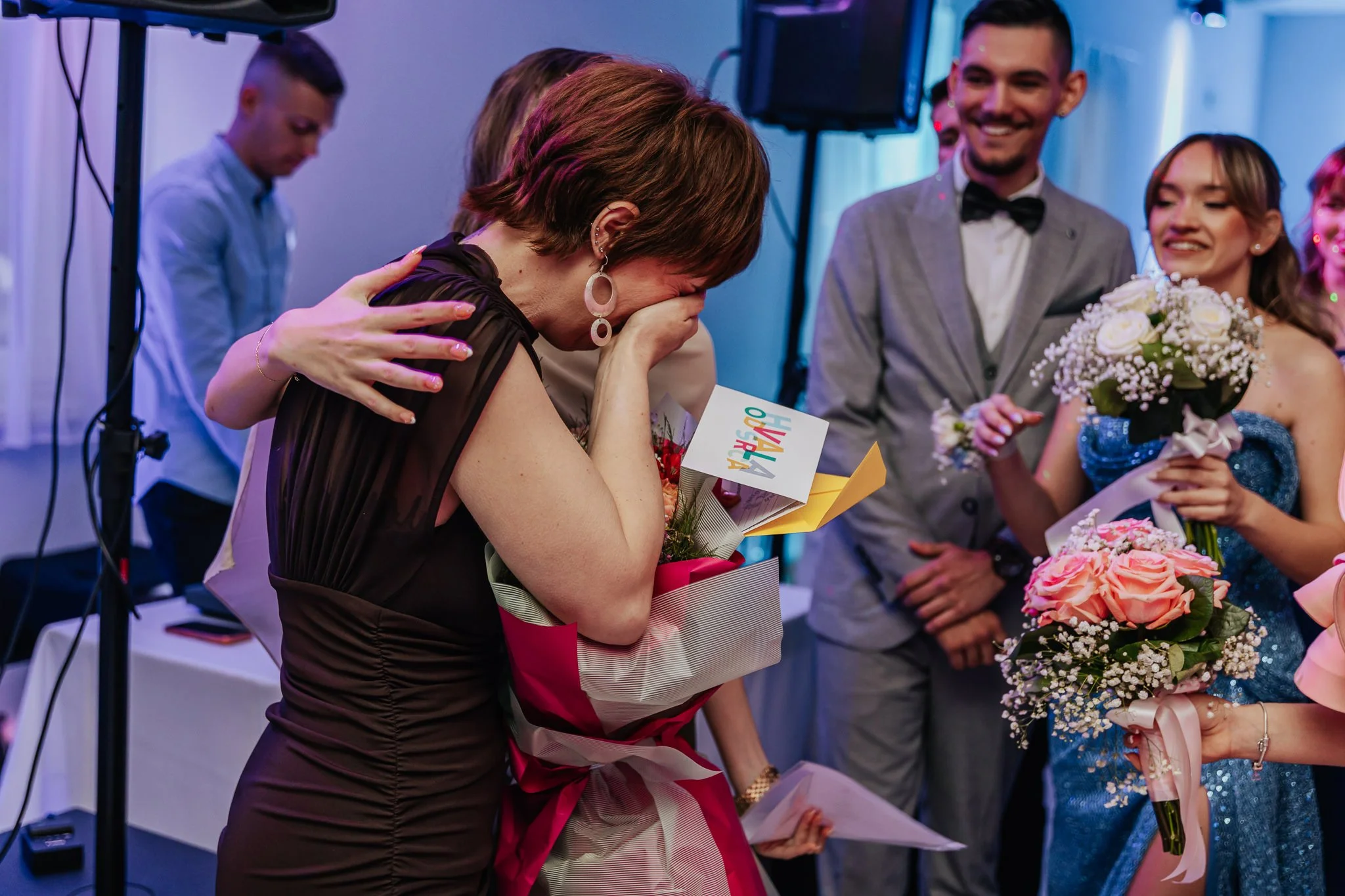 A grieving woman dressed in black is hugging a woman holding a bouquet of flowers at a celebration with a group of smiling people, including a woman in a blue dress holding flowers and a man in a grey suit with a bow tie, in a brightly lit indoor set