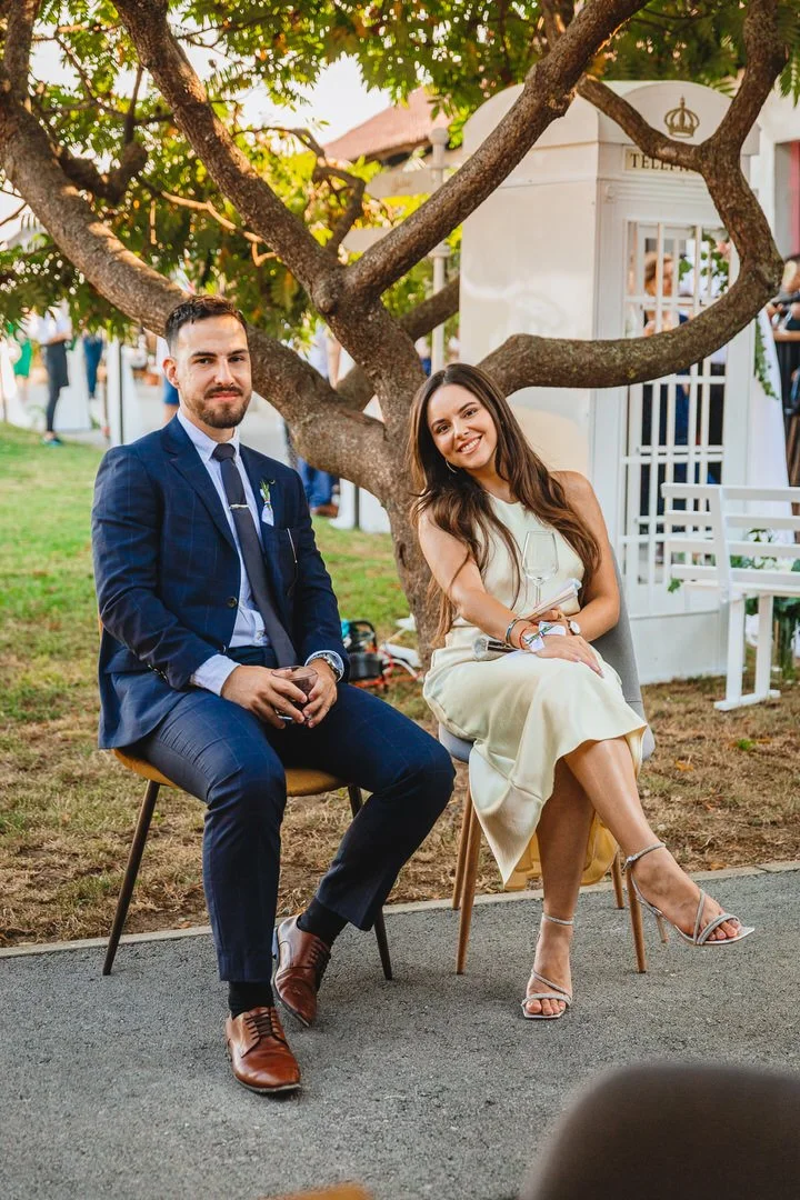 A man and a woman sitting outdoors on chairs under a large tree, with a white structure in the background. The man is wearing a blue suit, and the woman is in a light-colored dress. They are smiling.