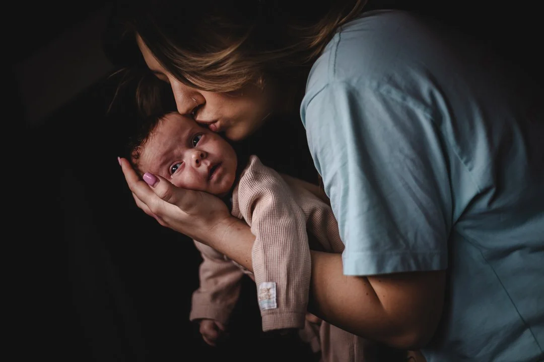 A woman gently holds a sleepy baby close, giving a kiss on the forehead in a dark setting.