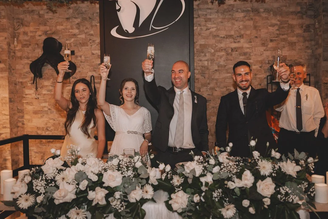 Group of six people celebrating with champagne at a wedding reception, standing behind a decorated table with white flowers and candles, in a brick-walled venue.