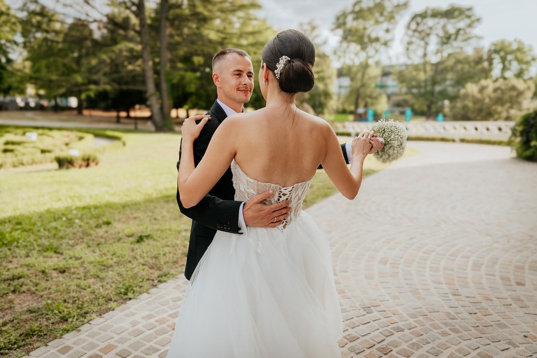 A newlywed couple dancing outdoors in a park in Našice, with the groom in a black suit and the bride in a white wedding gown holding a bouquet.