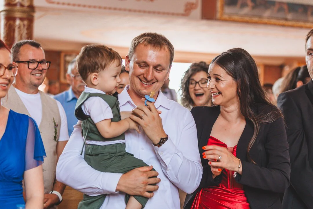 A group of people at a celebration, with a man holding a young boy and smiling, surrounded by smiling women and men in a decorated indoor setting.
