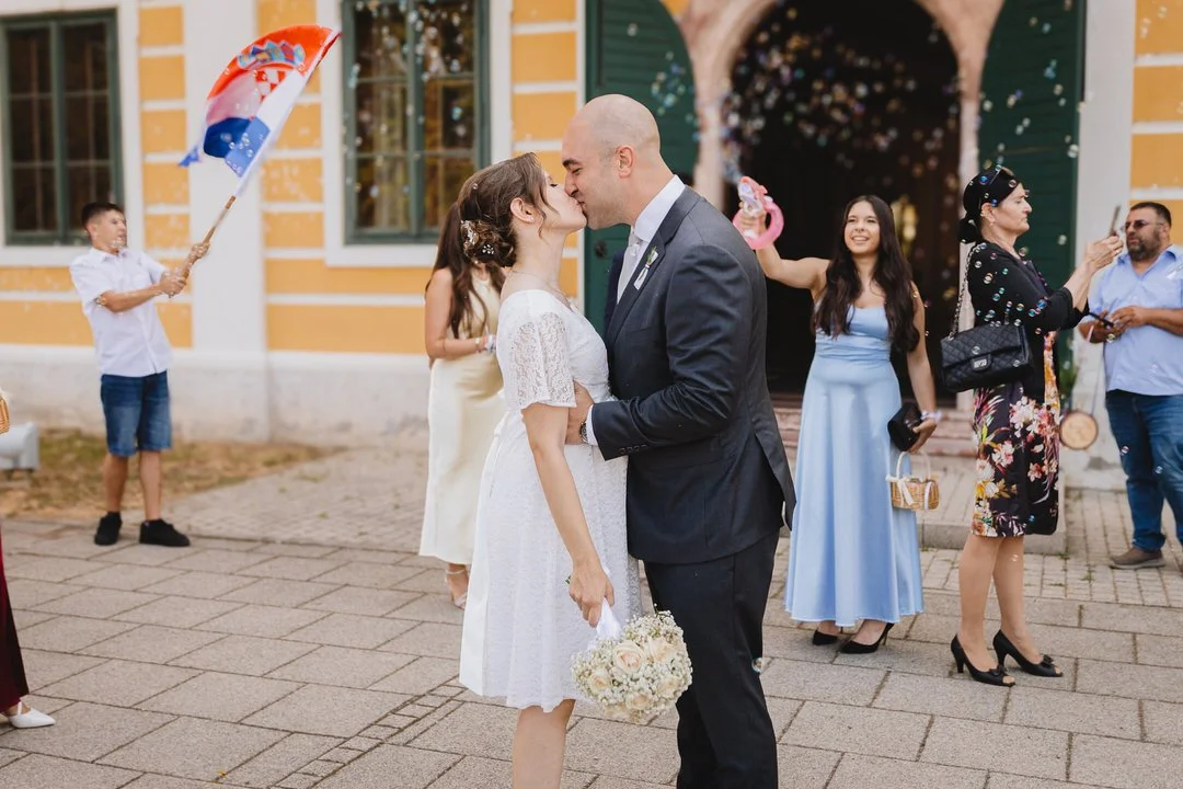 A newlywed couple shares a kiss outside a colorful building as friends celebrate around them with bubbles, an umbrella, and a phone photo, on a brick-paved street.