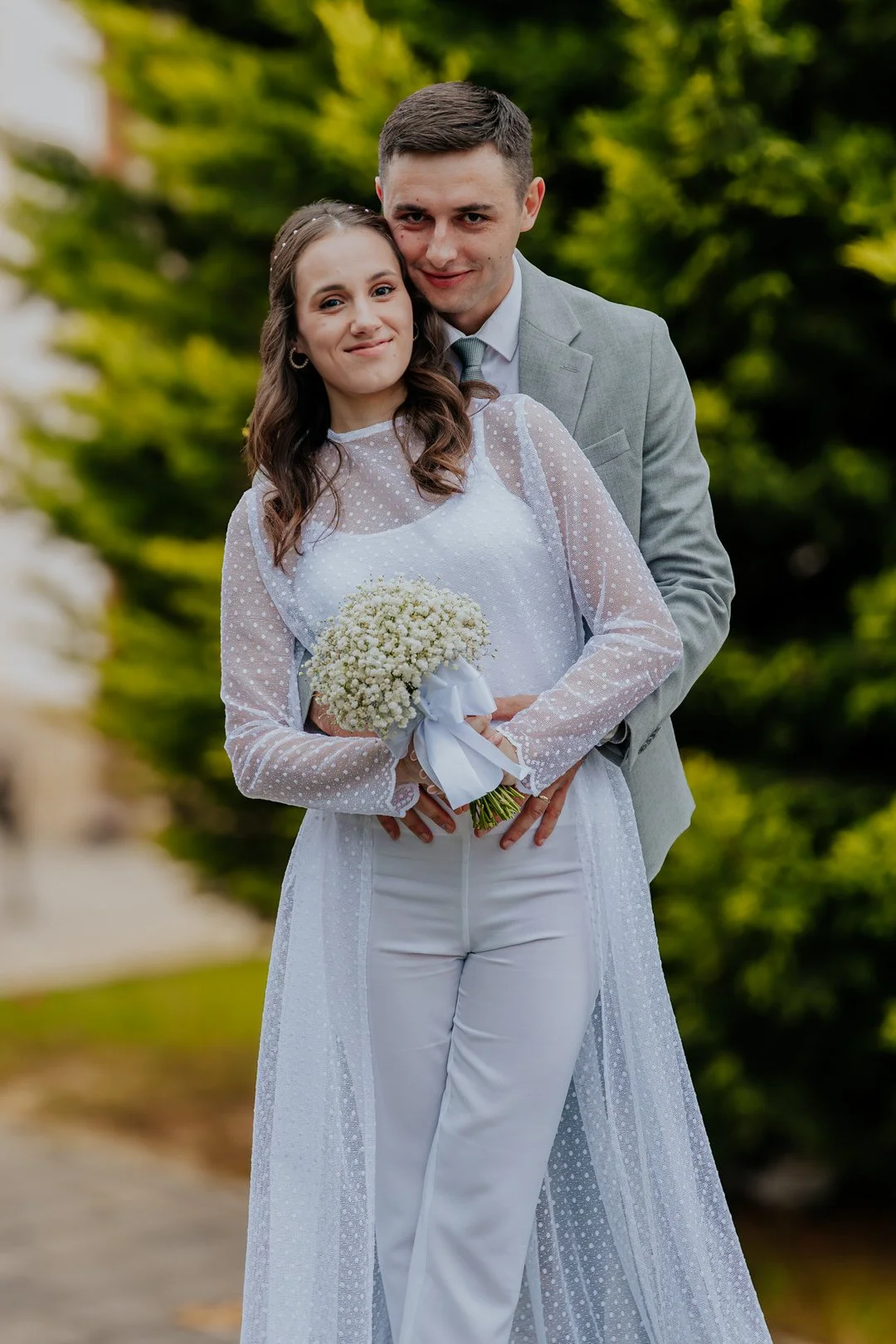 A couple dressed in wedding attire standing outdoors in front of green foliage, with the woman holding a bouquet of white flowers.