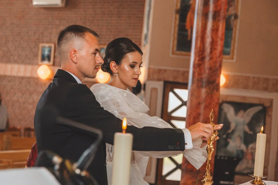 A bride and groom performing a wedding ceremony in a church in Jelisavac, with candles on the altar and religious artwork in the background.
