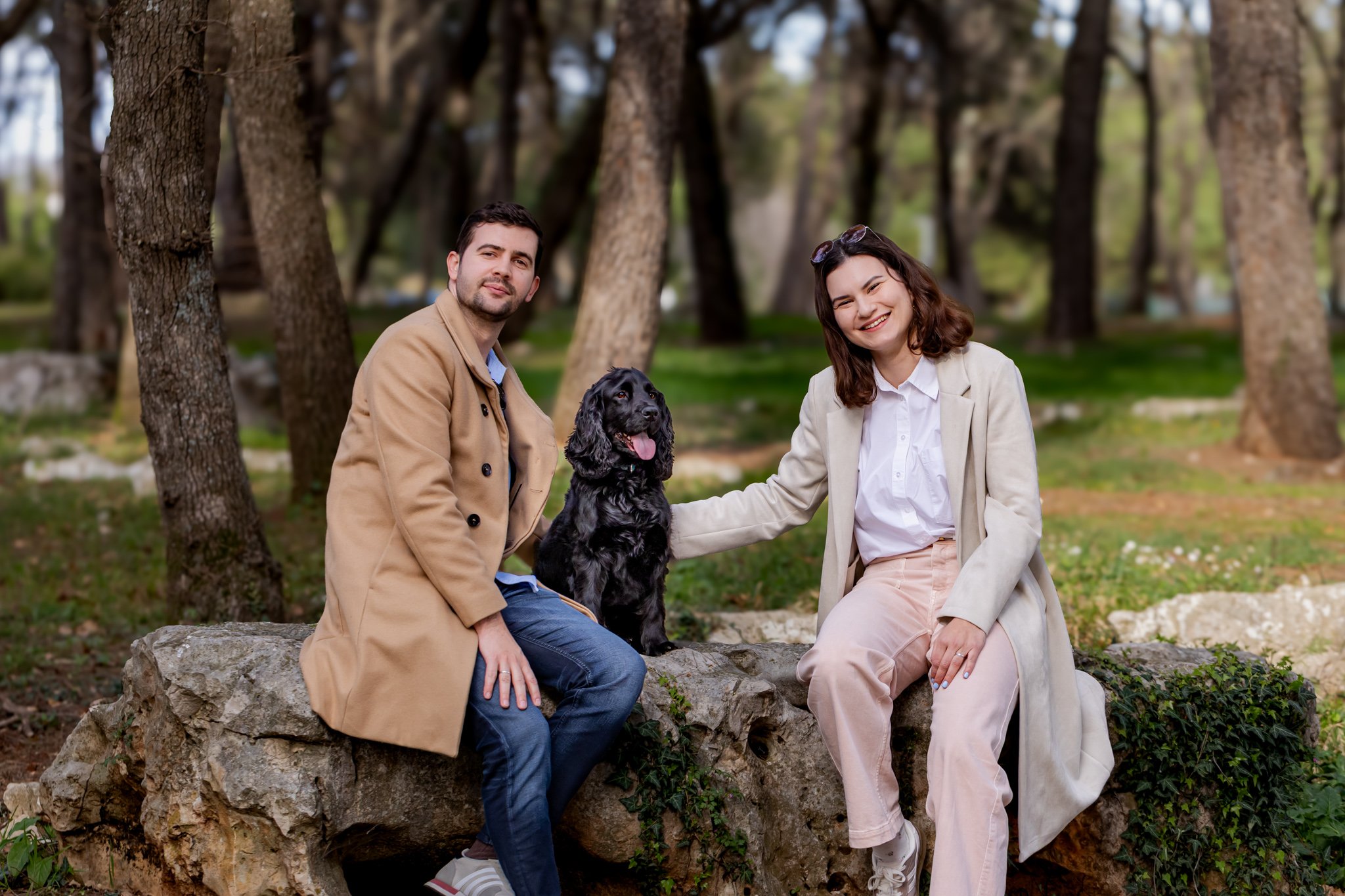 A man and woman sitting on a rock with a black English Cocker Spaniel in a wooded park during daytime in Umag Istria, Croatia.