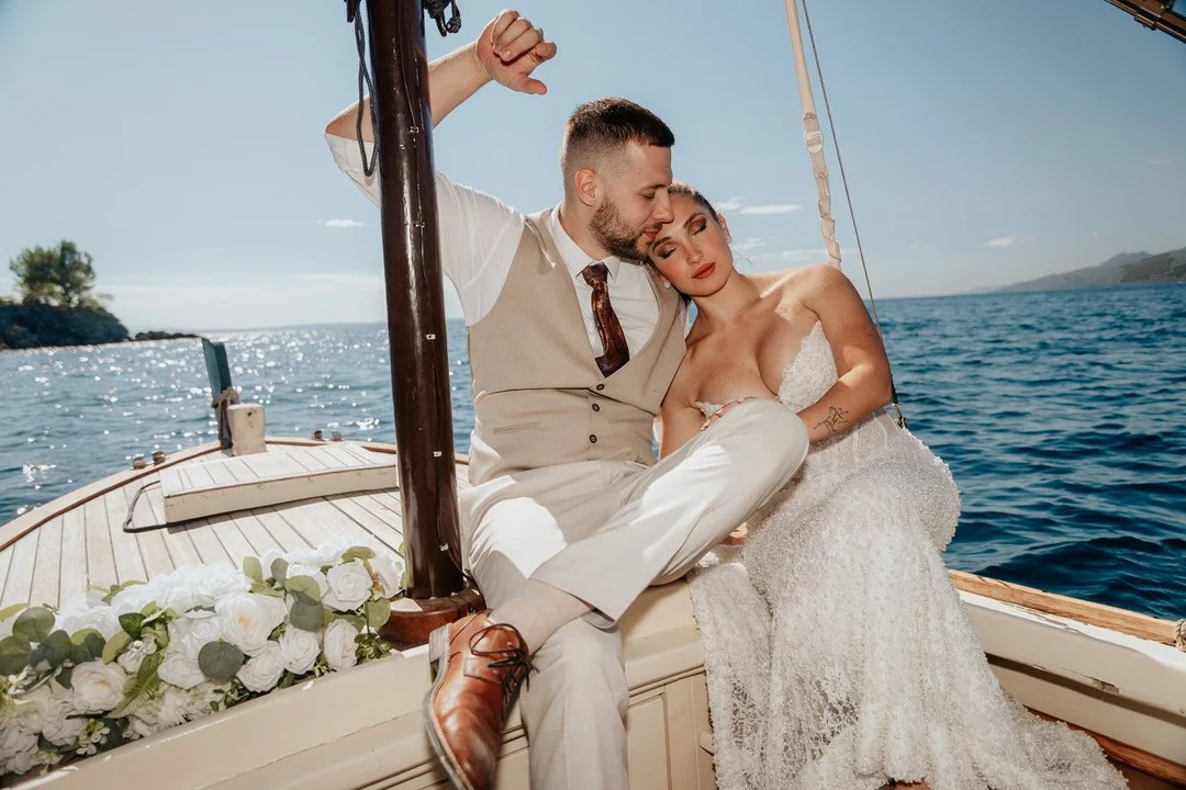 A newlywed couple sits on a boat with water and a distant shoreline in the background. The bride is wearing a white wedding dress and the groom is in a beige vest and trousers. They are embracing, with the groom resting his forehead on the bride's he