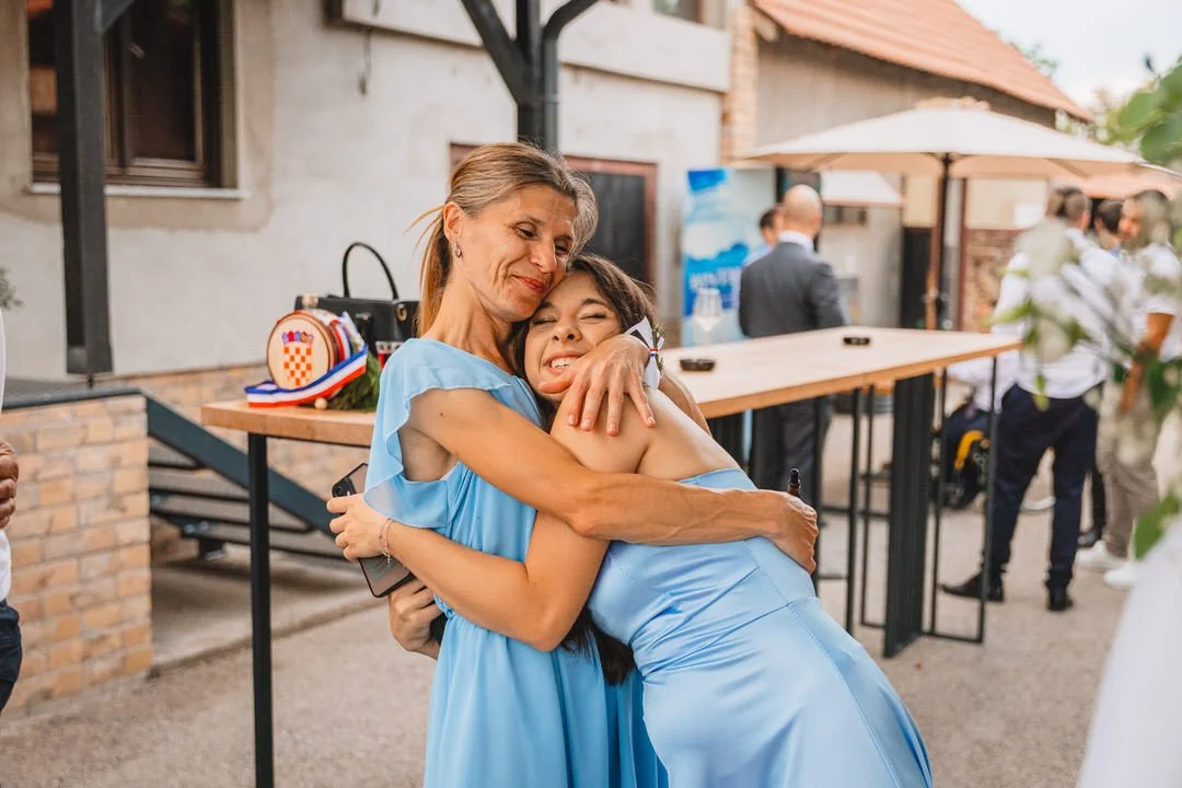 An older woman and a young woman hugging each other, both wearing light blue dresses, at an outdoor gathering or celebration.
