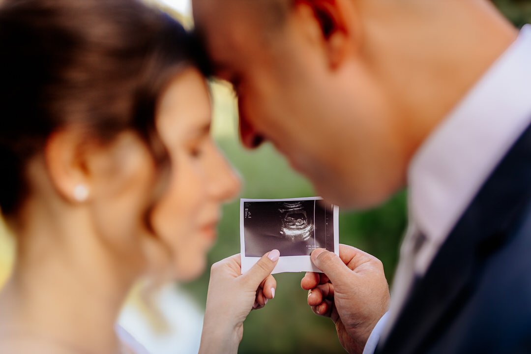 A couple holding a sonogram photo, with their foreheads touching, outdoors in a romantic moment.
