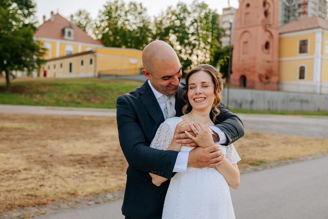 A man and woman embrace outdoors with historic buildings and trees in the background. The woman is wearing a white dress, and the man is in a dark suit. They are smiling happily.