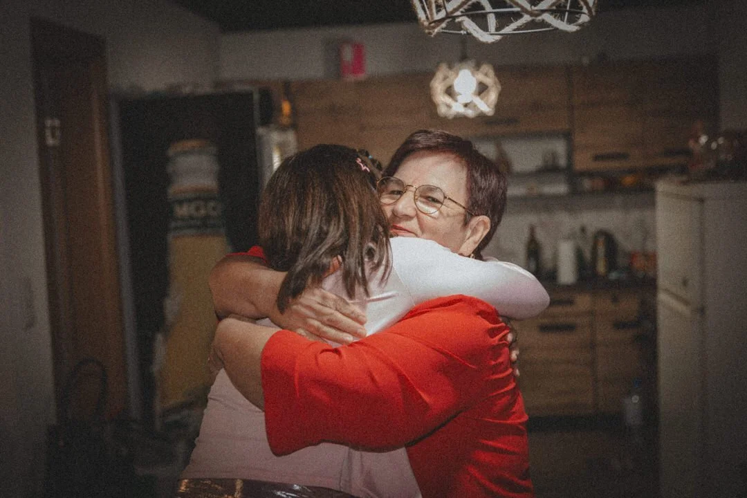 Two women hugging in a warmly lit kitchen, one wearing a red top and glasses, the other with short hair, sharing a joyful moment.