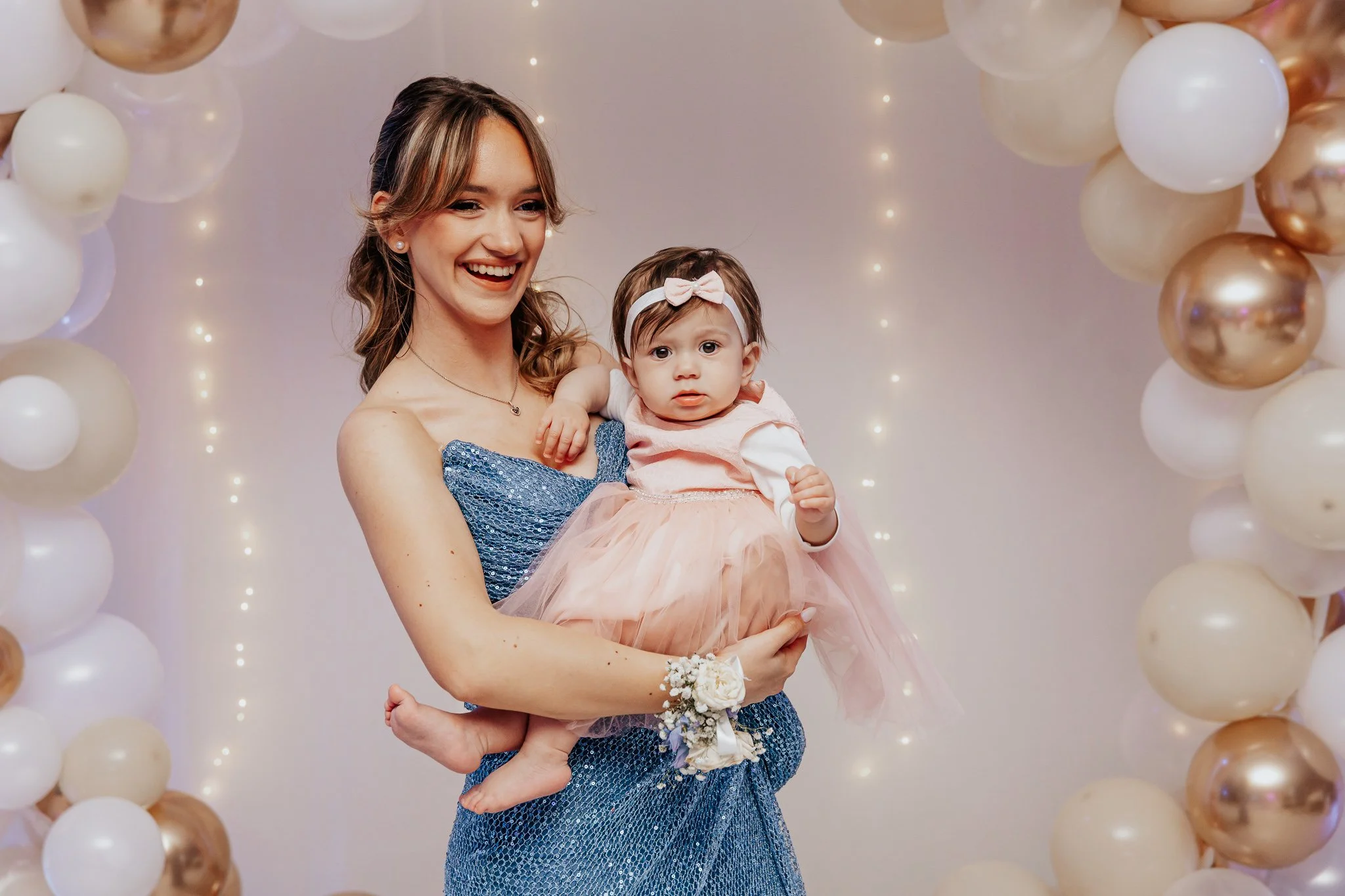 A woman smiling while holding a young girl in front of a backdrop decorated with white and gold balloons.