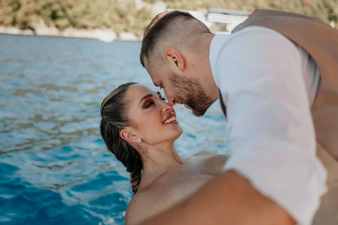 A couple is sharing an intimate moment by the water, with their foreheads touching and smiling at each other.