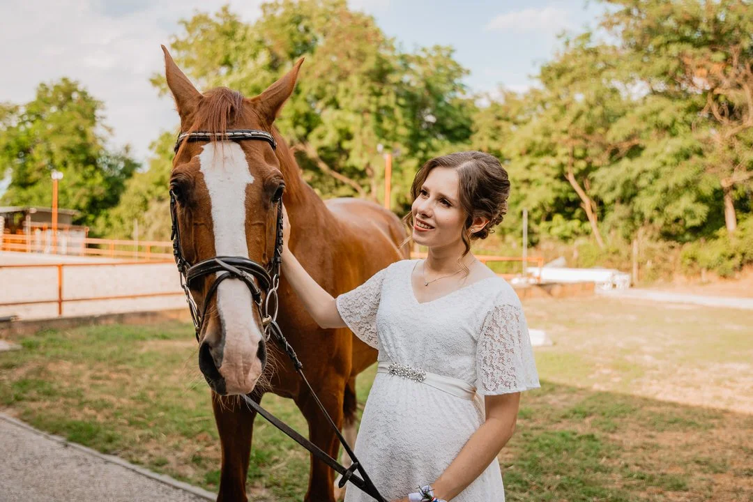 Young woman in a white lace dress smiling while petting a brown horse outdoors at Dunavski Raj wedding venue, with trees and a wooden fence in the background.