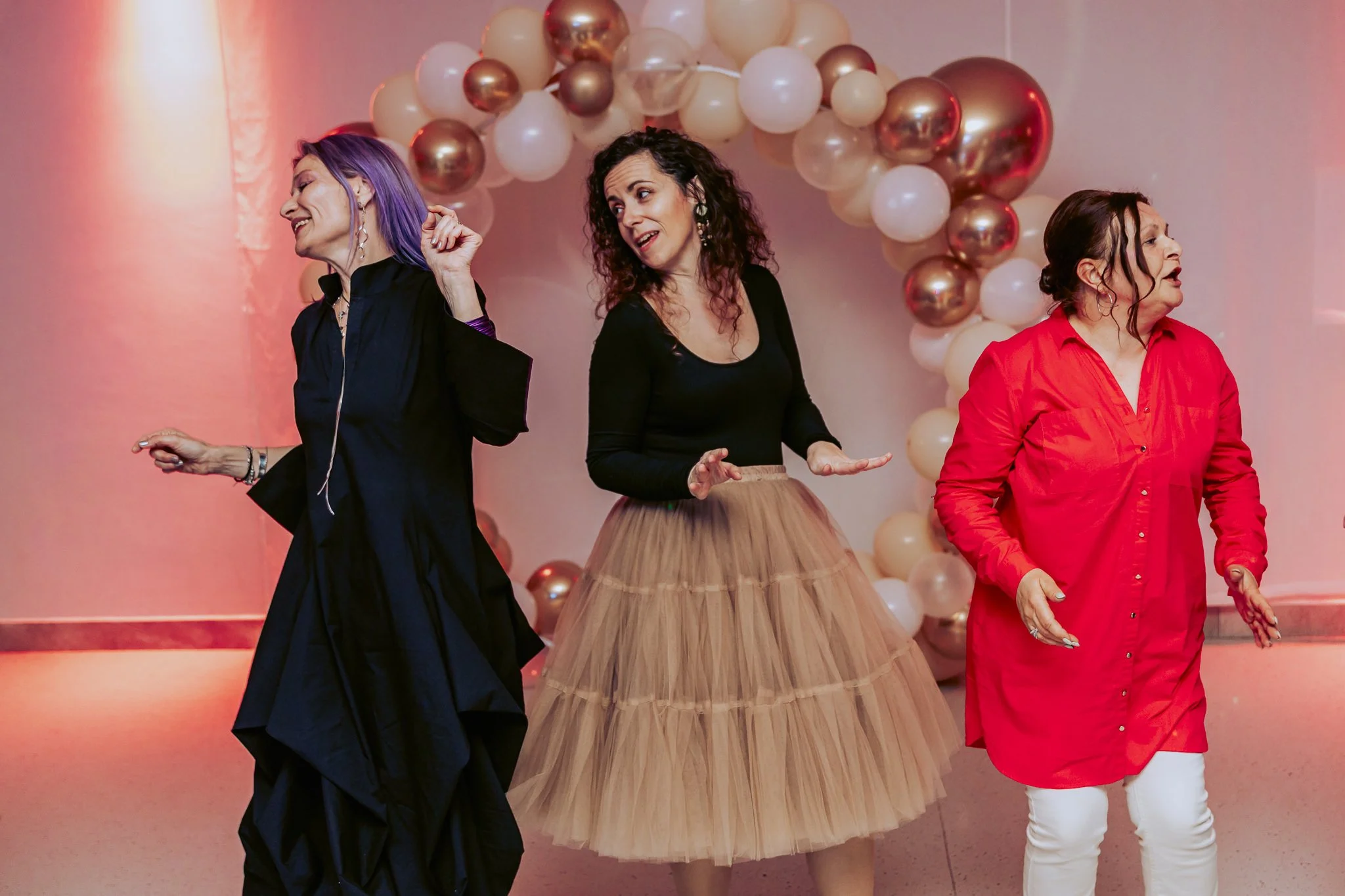 Three women dancing in front of a balloon arch with metallic gold, champagne, and white balloons, in a room with pink lighting.